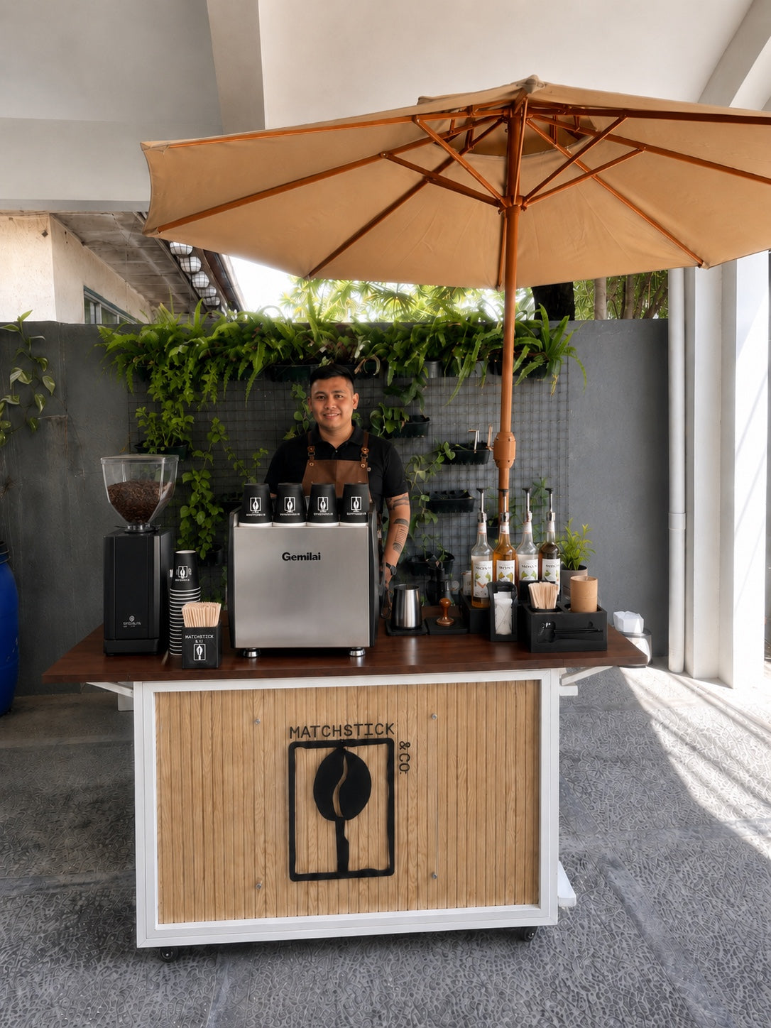 Matchstick & Co. coffee cart with a barista, espresso machine, and tan umbrella at an event.